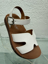 Cargar imagen en el visor de la galería, A single children's wedge sandal with white textured straps and a brown footbed, displayed on a clear acrylic stand against a textured white wall.
