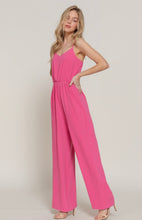 Load image into Gallery viewer, A woman modeling a bright pink sleeveless jumpsuit with a V-neckline and wide legs, standing against a plain light beige studio background.
